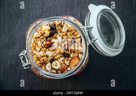 Konzept von hausgemachtem, handgemachtem geröstetem Müsli mit verschiedenen Nüssen und Rosinen in einem wunderschönen Glasgefäß, über der Aussicht. Schwarzer Textilhintergrund mit Platz. Stockfoto
