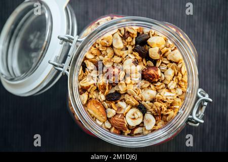 Konzept von hausgemachtem, handgemachtem geröstetem Müsli mit verschiedenen Nüssen und Rosinen in einem wunderschönen Glasgefäß, über der Aussicht. Schwarzer Textilhintergrund mit Platz. Stockfoto