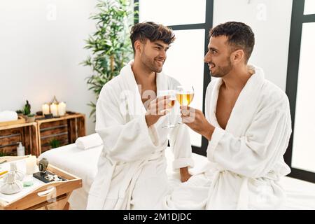 Zwei hispanische Männer toasten mit Champagner und sitzen auf dem Massageliegen im Beauty Center Stockfoto