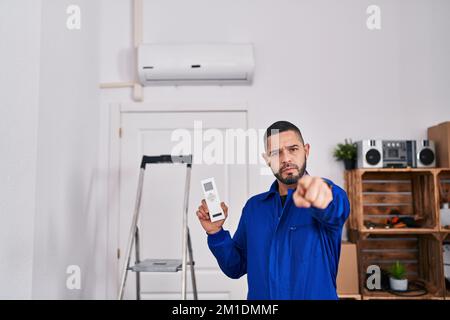 Hispanischer Servicetechniker, der mit der Klimaanlage arbeitet und mit dem Finger auf die Kamera und auf Sie zeigt, selbstbewusste Geste, die ernst aussieht Stockfoto