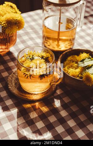 Der gesunde Tee aus der Teekessel und die Tasse aus Glas auf dem Tisch. Köstlicher Kräutertee aus frischen Dandelionblüten zu Hause am Sommertag. Grüne Clearing Heißer Deleliontee in einer Glas-Teekane. Bouquet von Nelkenkerblüten. Wildblumen Stockfoto