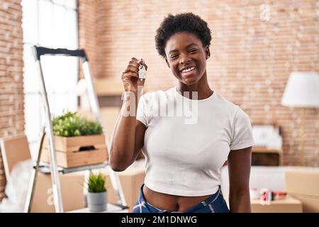 Eine afroamerikanische Frau, die die Schlüssel des neuen Heims hält und positiv und glücklich aussieht und mit einem selbstbewussten Lächeln lächelt und Zähne zeigt Stockfoto