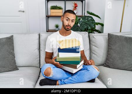 Ein junger afroamerikanischer Mann mit einem Stapel Bücher, der auf dem Sofa saß und müde aussah, erschöpft nach Müdigkeit und Kater, faule Augen in der Stockfoto