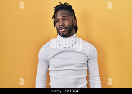 Ein afrikanischer Mann mit Dreadlocks, der einen Rollkragenpullover auf gelbem Hintergrund trägt, der zur Seite lächelt und wegstarrt und denkt. Stockfoto