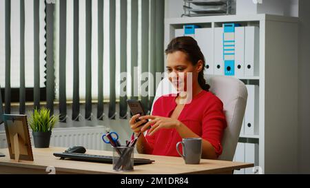 Hispanische Frau, die sich mit dem Smartphone im Büro unterhält. Unternehmer, der in einem beruflichen Arbeitsplatz arbeitet, Arbeitsplatz in einem privaten Unternehmen, der vor dem Computer auf dem Mobiltelefon tippt Stockfoto