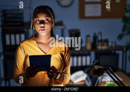 Eine afroamerikanische Frau mit Zöpfen, die nachts im Büro arbeitet, mit einem Tablet, das Fischgesicht mit Lippen macht, verrückte und komische Geste. Komischer Ausdruck. Stockfoto