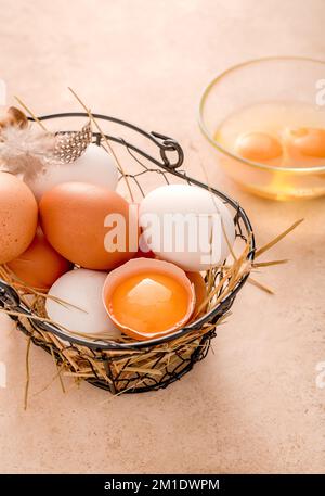 Verschiedene Eier im Metallkorb auf beigefarbenem Hintergrund. Rohe zerbrochene Eier in einer Glasschüssel. Stockfoto