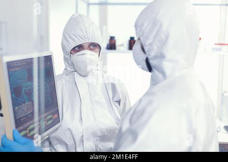 Wissenschaftler Kollegen im Overall diskutieren über die Virusentwicklung in ausgestatteten Laboren, die auf Computer-Desktops zeigen. Ärzte untersuchen Virusentwicklung mit High-Tech-Forschungsdiagnose gegen covid19 Stockfoto