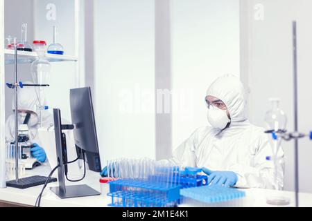 Mikrobiologe gekleidet in sterilem psa-Anzug, der nach Covid-Impfstoff forscht und auf pc-Bildschirm schaut. Mediziningenieur mit Computer während einer globalen Pandemie mit Coronavirus in Overall gekleidet. Stockfoto