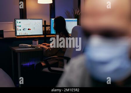 Geschäftsfrau trägt chirurgische Maske im Büro mit Kollegen. Gestresster Mann in der Gesellschaft arbeitet hart, um ein Projekt mit Gesichtsmaske als Sicherheitsvorkehrung aufgrund der Coronavirus-Pandemie zu beenden. Stockfoto