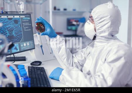 Medizinwissenschaftler hält Petrischale studiert Virusprobe gekleidet in bakteriologischen Anzug Ärzte analysieren Impfstoffentwicklung mit High-Tech-Forschung Diagnose gegen covid19 Virus. Stockfoto