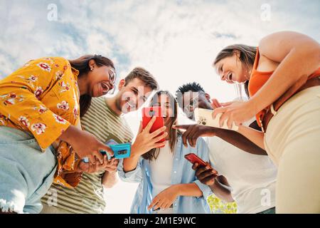 Niedrige Betrachtungswinkel einer Gruppe von lächelnden, multiethnischen Teenagern, die von Smartphones abhängig sind, lustige Videos anschauen, online einkaufen und draußen Spaß haben. Multiethnische, fröhliche junge Leute, die auf der Suche sind, mit verschiedenen Anwendungen auf ihren Handys unterhaltsam unter blauem Himmel. Verbindungskonzept. Hochwertiges Foto Stockfoto