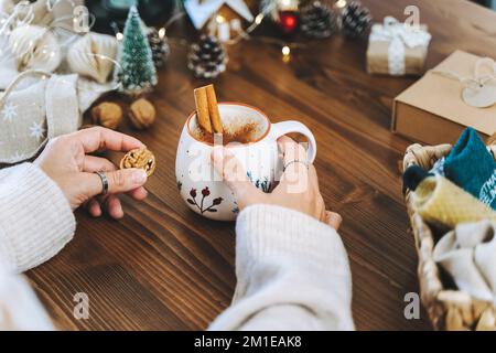 Weihnachtsbecher Kaffee mit Zimt in Frauenhänden. Geschenkverpackung für Weihnachtsgeschenke, Nahaufnahme. Vorbereitung der Geschenke auf einem Holztisch mit natürlicher Einrichtung Stockfoto