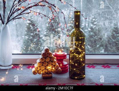 Weihnachtsdekoration Set mit Weinflasche gefüllt mit Mikro-LED-Partylichtern und Fichte aus gebrauchten Weinkorken, dahinter ist ein Fenster mit Schnee. Stockfoto