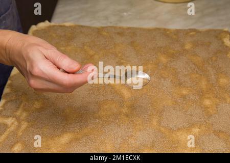 Gerollter Teig mit Zucker und Zimt für Backbrötchen. Zucker mit Zimt in Löffel Stockfoto