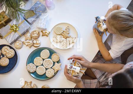 Das kleine süße Mädchen und der Junge spielen mit gestrickten Schneemännern, essen Lebkuchen und trinken Kakao mit Marshmallows. Stockfoto