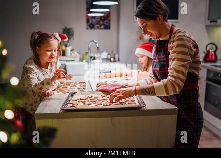 Die Schwestern bereiten Weihnachtskekse zu, während Mutter aus selbstgemachtem Teig Lebkuchen zusammen ein Backblatt in der Küche auslegt. Einen schönen Familienurlaub Stockfoto