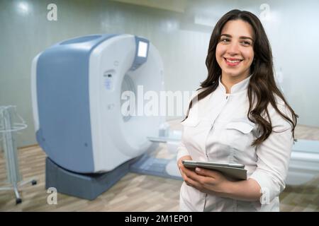 Arzt mit einem digitalen Tablet, der lächelt und auf die Kamera im Hintergrund eines CT-Scanners schaut Stockfoto