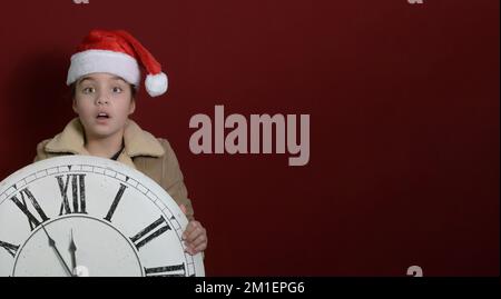 Ein Mädchen mit einer großen, alten Wanduhr in der Hand. Er trägt einen Weihnachtsmannmütze, einen begeisterten Blick in die Kamera. Platz für Text. Fünf Minuten bis zum neuen Jahr Stockfoto