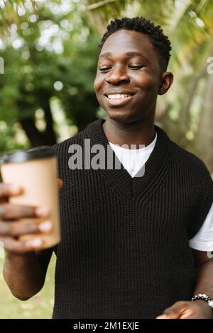 Ein junger afrikanischer Mann lächelt, um draußen eine Kaffeetasse zu trinken. Junger schwarzer Student in Kaffeepause im Park Stockfoto