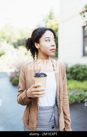 Junge afrikanische multirassische Frau hält Einweg braun Papier Kaffeetasse auf der Straße im Freien. Gesichtsprofilaufnahme, Seitenansicht im Hochformat. Kaffee oder Tee zum trinken Stockfoto