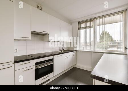Eine Küche mit weißen Schränken und schwarzen Ablageflächen auf der Insel vor dem Waschbecken ist neben dem Fenster Stockfoto