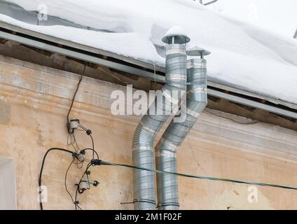 Zwei Abluftrohre aus Metall an der Wand eines alten Hauses. Das Dach des Gebäudes in weißem Schnee. Bewölkt, kalter Wintertag, weiches Licht. Stockfoto