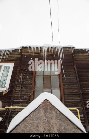 Eis, mehrere Eiszapfen hängen am Dachrand, im Winter oder Frühling. Plankenwand eines alten Holzhauses mit Fenstern. Große Eiszapfen in glatten, schönen Reihen. Bewölkter Wintertag, weiches Licht Stockfoto