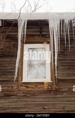 Lange, gefährliche Eiszapfen hängen an der Dachkante, im Winter oder Frühling. Holzwand eines alten Holzhauses mit Fenstern. Große Eiszapfen in glatten, schönen Reihen. Bewölkter Wintertag, weiches Licht Stockfoto