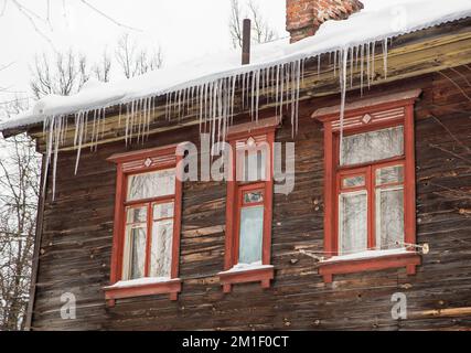 Vereiste, lange Eiszapfen hängen am Dachrand, im Winter oder im Frühling. Plankenwand eines alten Holzhauses mit Fenstern. Große Eiszapfen in glatten, schönen Reihen. Bewölkter Wintertag, weiches Licht. Stockfoto