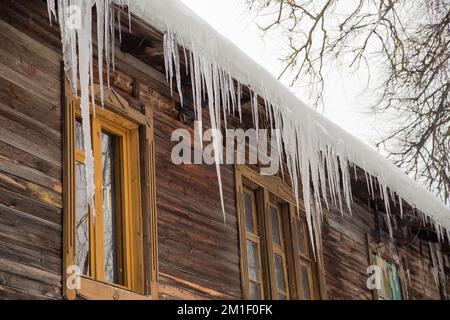 Scharfe Eiszapfen hängen im Winter oder Frühling am Dachrand. Holzwand eines alten Holzhauses mit Fenstern. Große Eiszapfen in glatten, schönen Reihen. Bewölkter Wintertag, weiches Licht. Stockfoto