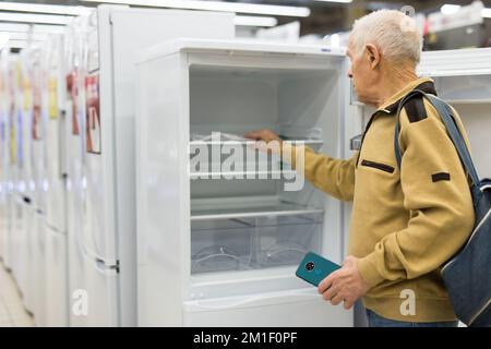 Älterer grauhaariger Rentner, der am Schalter im Ausstellungsraum der Abteilung für Elektrogeräte einen Kühlschrank sah Stockfoto