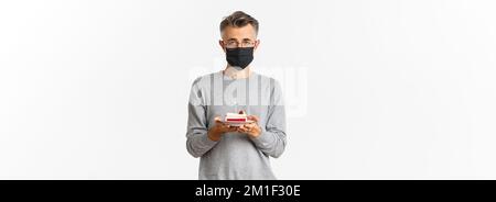 Konzept von Coronavirus, Quarantäne und Urlaub. Bild von traurigen Mann mittleren Alters in schwarzer medizinischer Maske und Brille, hält Geburtstagskuchen mit brennenden Kerze Stockfoto