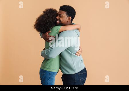 Schwarzer Mann und Frau, die ihr Glück ausdrücken und sich isoliert in beigefarbenem Hintergrund umarmen Stockfoto