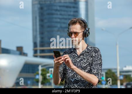 Ein junger Mann mit einem Smartphone in Kopfhörern und Sonnenbrillen hört Musik im Hintergrund moderner Gebäude Stockfoto