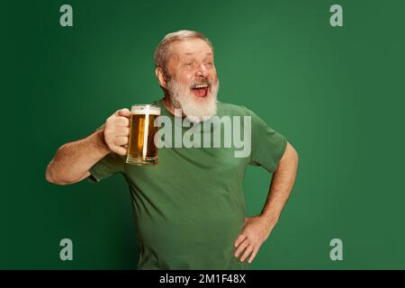 Porträt eines Seniorenmannes in T-Shirt-Pose mit Bier isoliert auf grünem Hintergrund. St. Patrick's Day-Feier. Betrunkener Blick Stockfoto