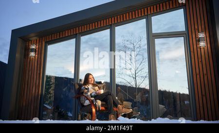 Junge Frau, die auf der Terrasse des modernen Scheunenhauses in den Bergen ruht. Glückliche weibliche Tourist mit einer Tasse Tee, genießen Winterurlaub in einem neuen Cottage. Stockfoto