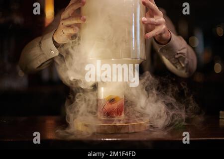 Ein köstlicher Negroni-Rauchercocktail in einem Glas, den der Barkeeper mit einem Rauchglas bedeckt Stockfoto