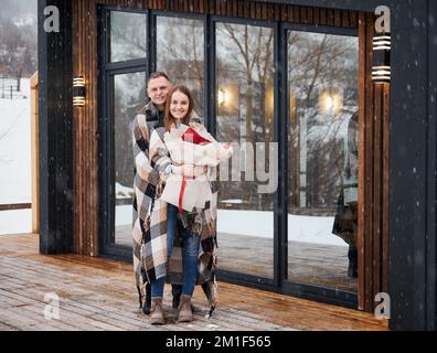 Ein glückliches Paar in voller Länge, das vor einem skandinavischen Haus steht, unter wunderschönem Schnee. Ein Mann mit einer Decke, umarmte die Freundin und lächelte, während eine Frau einen Rosenstrauß hielt. Stockfoto