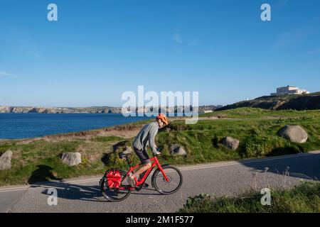 Ein Radfahrer mit Schutzhelm, der auf einem roten Trek-Fahrrad auf der Küstenstraße mit Blick auf Newquay Bay in Cornwall in Großbritannien bergauf fährt. Stockfoto