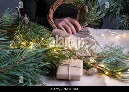 Ich mache einen rustikalen Weihnachtskranz. Weibliche Hände auf dem Hintergrund eines Holztischs mit Kiefernästen, Kegeln, Lichtern. Feiertagsvorbereitungen Stockfoto