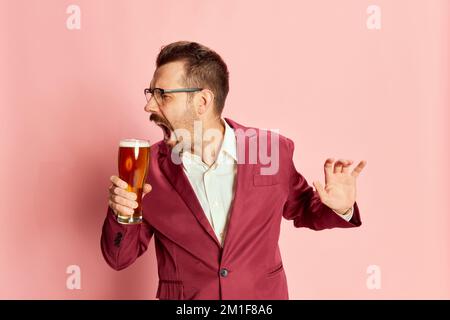 Porträt eines stilvollen, emotionalen Mannes in einem Anzug, der mit einem Glas Lagerbier auf pinkfarbenem Hintergrund posiert. Singen, trinken Stockfoto