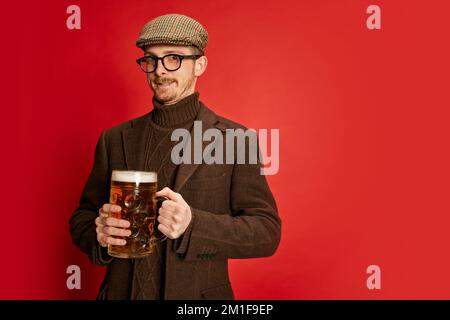 Porträt eines stilvollen Mannes in klassischer Kleidung, Mütze und Gläsern, der mit einem Glas schäumendem Bier auf rotem Hintergrund posiert. Bayerisch Stockfoto