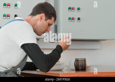 Mittagspause. Essen am Arbeitsplatz während der Arbeit. Ein weißer Mann im Arbeitsanzug sitzt am Tisch in der Produktionshalle und isst aus dem Container. Der Arbeiter isst im Pausenraum zu Mittag. Stockfoto