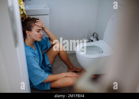 Junge kranke Frau, die auf dem Boden neben der Toilette sitzt. Stockfoto