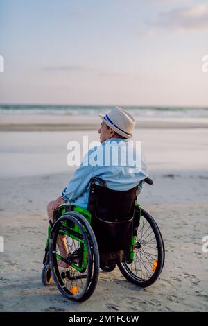 Rückblick auf einen Mann, der auf einem Rollstuhl sitzt und den Blick auf das Meer genießt. Stockfoto