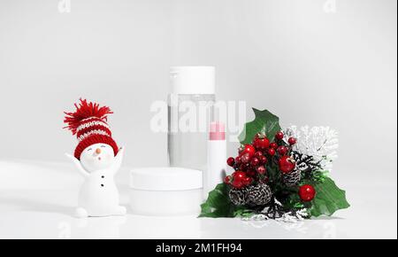 Hautpflegeflaschen auf weißer Oberfläche mit Schneemann. Kosmetik der Wintersaison. Leere Behälter mit Geschenken und Tannenzweigen im Hintergrund. Weihnachtsvorsteher Stockfoto