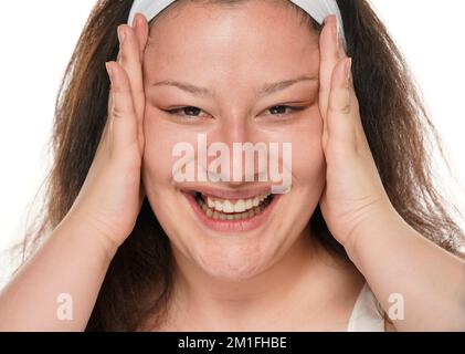 Junge, fröhliche, dicke Frau, die ihre Gesichtshaut mit ihren Handflächen auf weißem Hintergrund zusammenzieht. Stockfoto