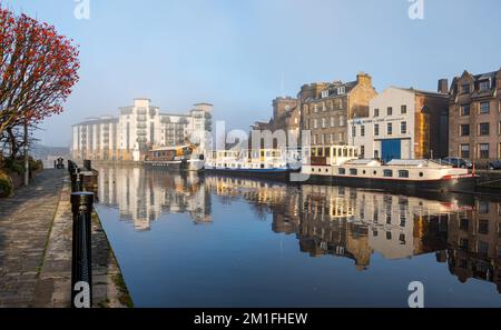 Leith, Edinburgh, Schottland, Großbritannien, 12.. Dezember 2022. Wetter im Vereinigten Königreich: Sonnenschein und Nebel. An einem bitterkalten Tag, an dem die Temperatur nicht über Null Grad steigt, Die Sonne scheint auf das Wasser von Leith und erzeugt farbenfrohe Reflexionen im Fluss mit eiskaltem Nebel, der die Aussicht in der Ferne eines modernen Blocks von Wohnungen und Hausbooten verdeckt, die sich im stillen Wasser am Flussufer spiegeln. Kredit: Sally Anderson/Alamy Live News Stockfoto