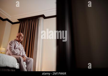 Trauriger, depressiver Senior-Mann im Satin-Pyjama, der auf dem Bett sitzt Stockfoto